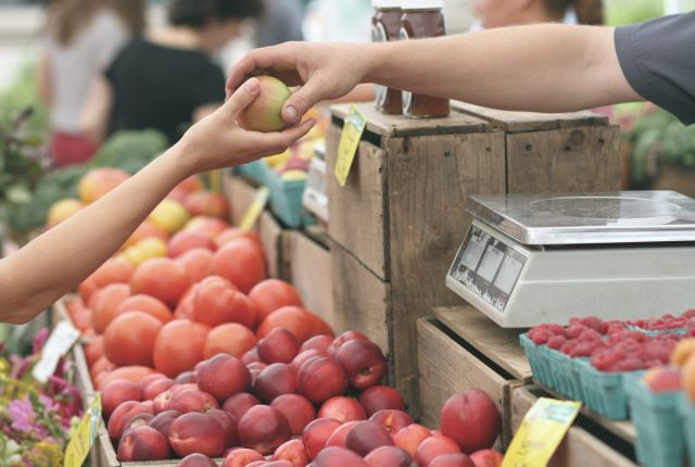 Zdjęcie przedstawia stoisko owocowe. Widoczne sa jedynie ręce klienta i sprzedawcy, które przekazują sobie jabłko.