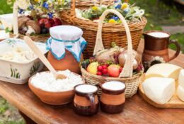 Sliced cheese, cottage cheese in a bowl, milk in jugs, fruits and vegetables in a basket, jam in a jar covered with a napkin. All on a wooden table in the open air